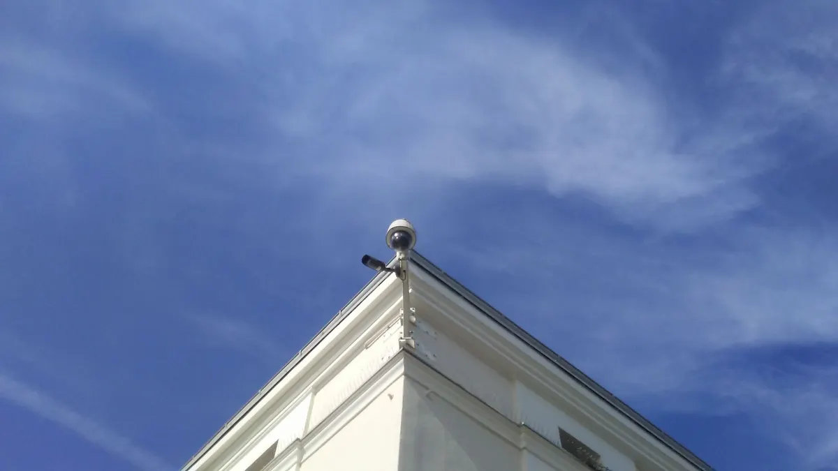 Câmera dome PTZ e câmera bullet presas à quina do telhado de um prédio branco, contra o céu azul com nuvens finas.