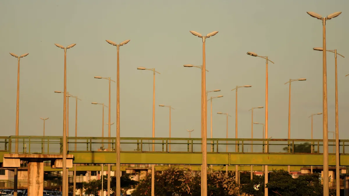 Dezenas de postes altos de iluminação pública com luminárias duplas ao pôr do sol, sobre uma passarela verde de pedestres e copas de árvores.