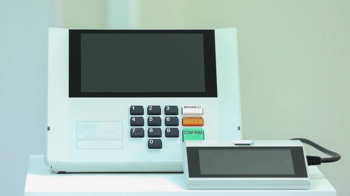 Urna eletrônica brasileira modelo UE2020 com o terminal do mesário ao lado, sobre uma mesa branca, contra um fundo claro.